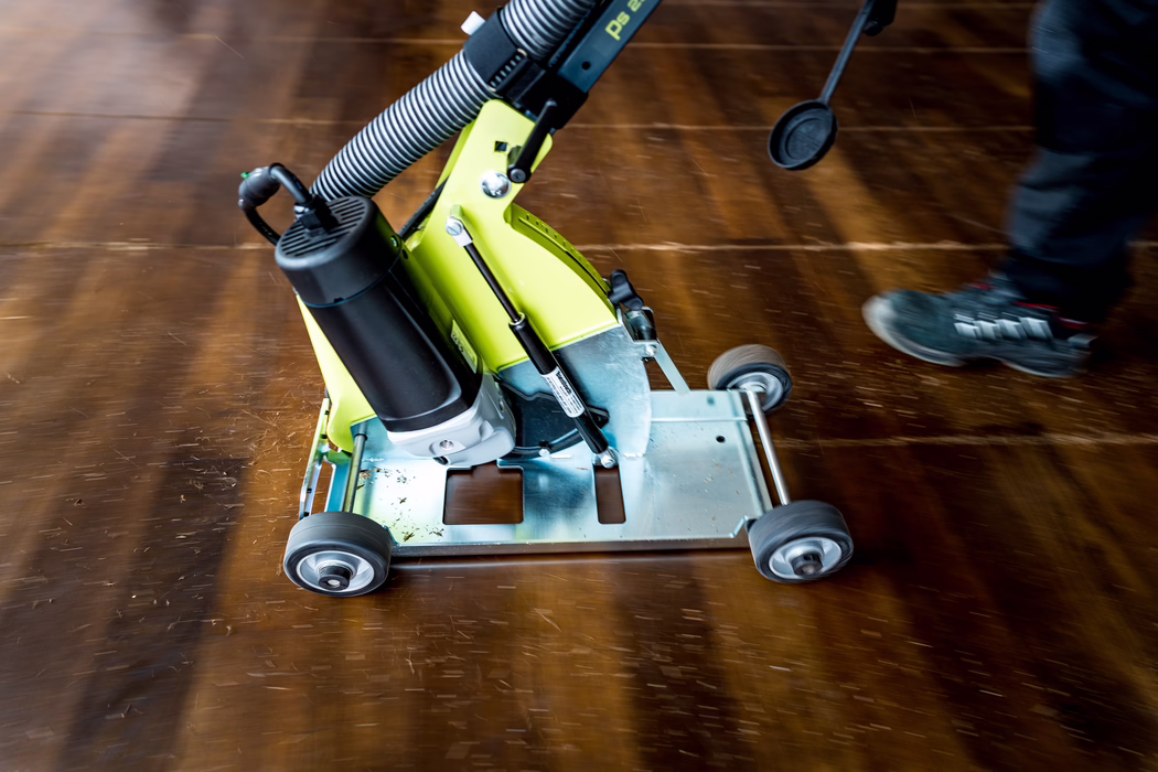 Close-up of a green floor sanding machine on a wooden floor; the machine's metal base with four small wheels is visible, along with dust and wood shavings. A person's lower leg wearing a sneaker appears at the right edge, indicating an operator. The wooden floor shows sanding marks and a warm brown tone.