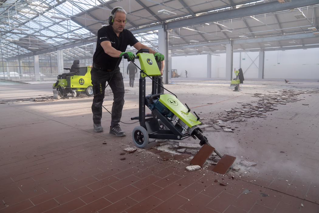 WOLFF_Application Images_RamHammer_Main_2022-11 A construction worker uses a green electric floor-removal machine to chip away ceramic tiles in a large, sunlit industrial hall. He wears a black polo shirt, green work gloves and hearing protection while pushing the wheeled machine over broken tiles and dust. In the background other equipment and workers are visible under a high glass roof; debris and loose tiles cover the floor. The scene depicts active floor renovation with dust, cables and safety gear.