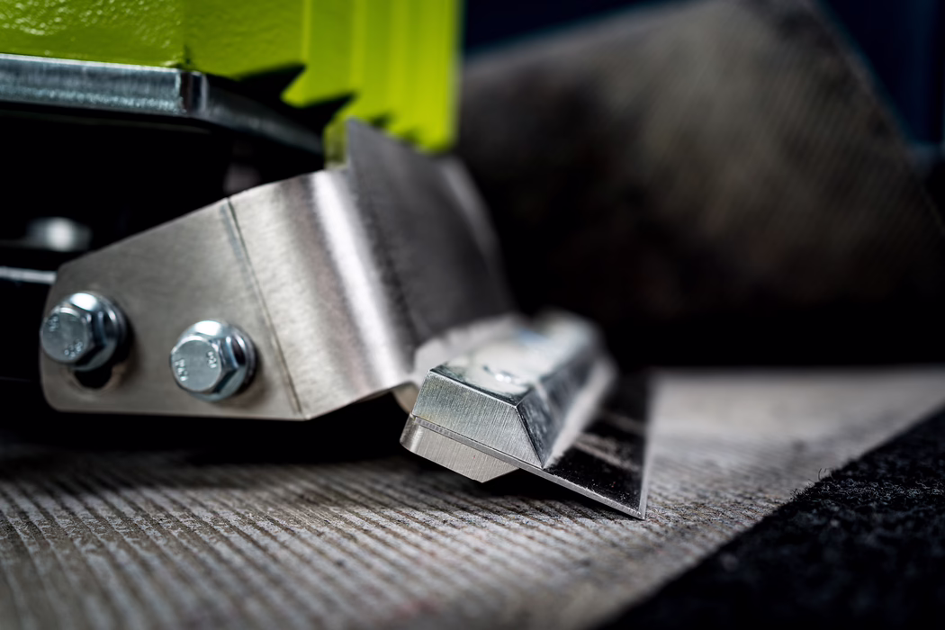 WOLFF_Application Image_Lupo_2024-04 (23) Macro close-up of a metallic floor tool: a beveled, sharpened steel blade mounted on a metal bracket with two bolts, attached to a bright yellow-green housing. The tool rests on textured flooring (carpet or underlay); the brushed steel, screw heads and folded metal show fine scratches and machining marks. A shallow depth of field isolates the blade in the foreground while the background fades into soft bokeh, emphasizing industrial detail, tactile surfaces and cool metallic tones. The image conveys precision, craftsmanship and the functional character of a flooring or carpet installation scraper.