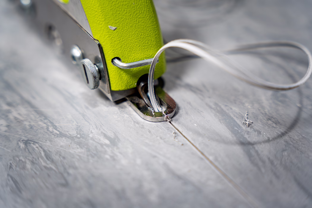WOLFF_Application Image_Groover_2024-01 (4) Macro close-up of the metal foot area of a small handheld sewing or fabric tool with a lime-green painted body. The image shows chrome hardware including a knurled adjustment screw and a polished metal guide plate through which a translucent thread or filament is looped. Tiny shavings and a fine seam line are visible on the textured grey work surface below. A shallow depth of field keeps the metal foot sharply in focus while the background and the looped thread blur into a soft bokeh, conveying a sense of careful, detailed workmanship and material texture.