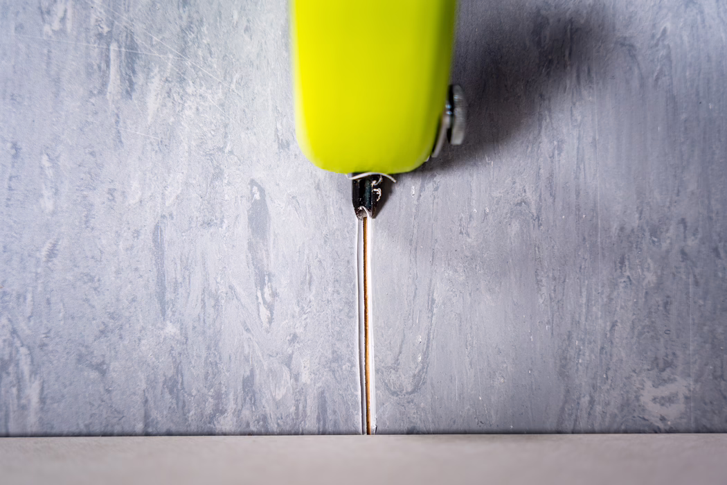 WOLFF_Application Image_Groover_2024-01 (12) Close-up view of a lime-green retractable utility knife positioned vertically above a narrow seam between a textured gray wall and a light-colored floor. The thin metal blade is extended downward into the seam, with the plastic handle and a small metal screw visible at the top of the frame. The image highlights contrasting textures — the rough, painted plaster of the wall and the smoother floor surface — and is softly lit from above, revealing subtle scratches and wear on the blade and handle.
