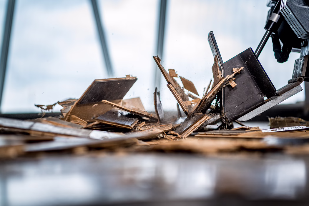 Close-up of a floor demolition: a power tool's metal scraper presses into layered wooden floorboards, breaking them into splintered fragments and dust. The shallow depth of field emphasizes sharp, jagged wood chips and fibers in the foreground while a soft, cool-toned windowed background is out of focus. Warm brown tones of the damaged parquet contrast with the muted blue-gray light, conveying dynamic texture and the sense of renovation or removal work.