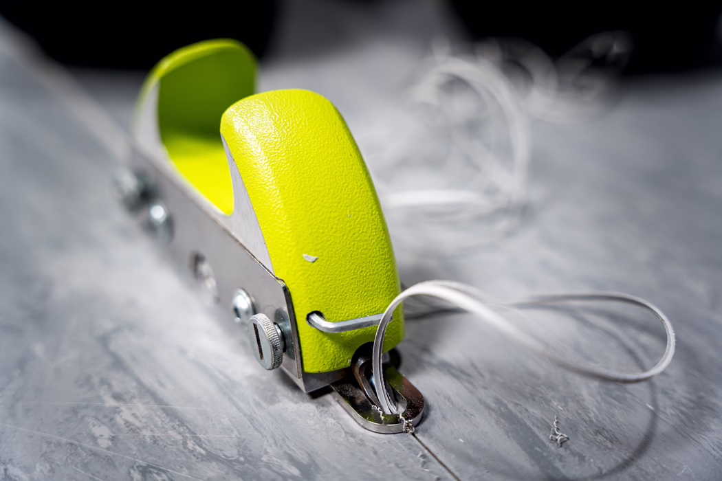 WOLFF_Application Image_Groover_2024-01 (5) Close-up of a small lime-green handheld single-hole punch tool resting on a gray marbled work surface. The textured plastic top has a bright chartreuse color with minor scuffs and a small paint chip; the metal body shows exposed screws, a knurled adjustment dial and a folded metal throat. A loop of translucent elastic cord is threaded through the front opening and lies coiled on the surface. The photograph uses a shallow depth of field, keeping the punch sharply in focus while the background is softly blurred, emphasizing details such as the grainy plastic texture, subtle chrome reflections and faint scratches on the tabletop.