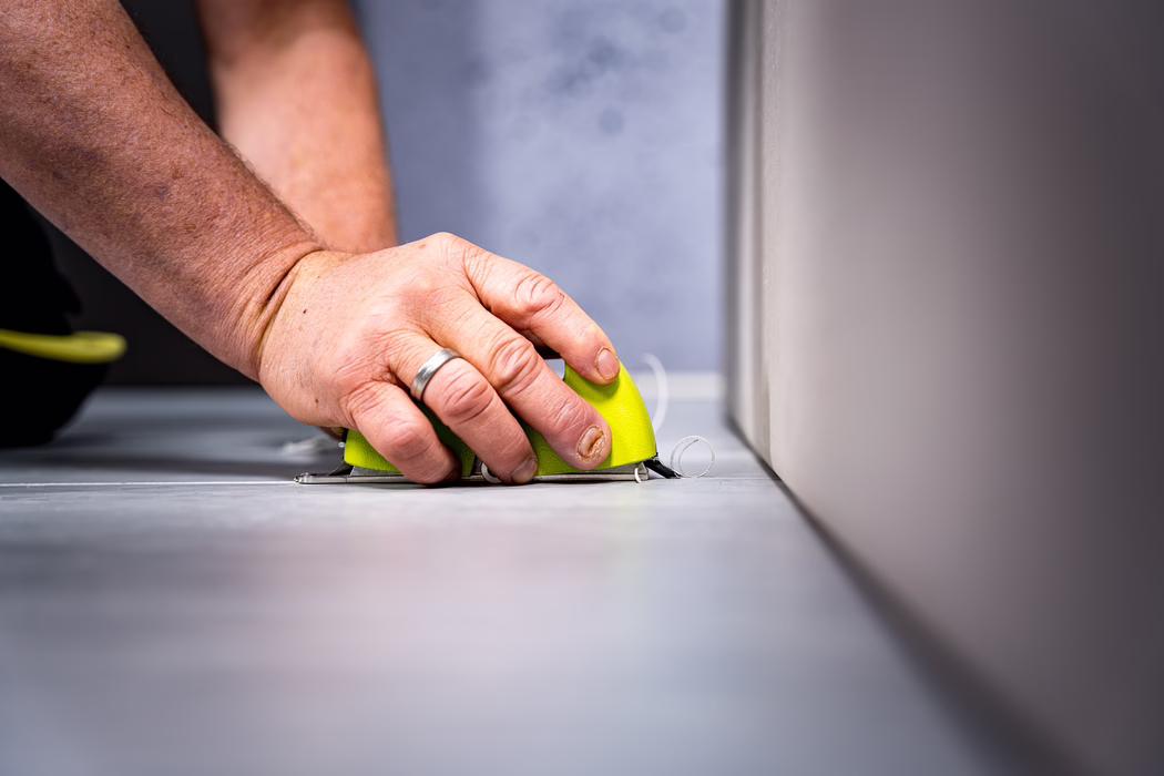 WOLFF_Application Image_Groover_2024-01 (9) A close-up photograph shows a person's hand holding a small lime-green floor trimming tool pressed against the seam between a smooth gray floor and a vertical wall. The hand wears a simple metal ring on the ring finger and has short, slightly worn fingernails; the skin shows natural texture and small blemishes. Thin, curled shavings of flooring material are visible at the tool's cutting edge as the operator carefully trims along the wall; the metal blade and the base of the tool are in sharp focus while the foreground and background are softly blurred, creating a shallow depth of field. The image conveys detailed workmanship, attention to a precise edge, and a quiet, professional indoor work setting.