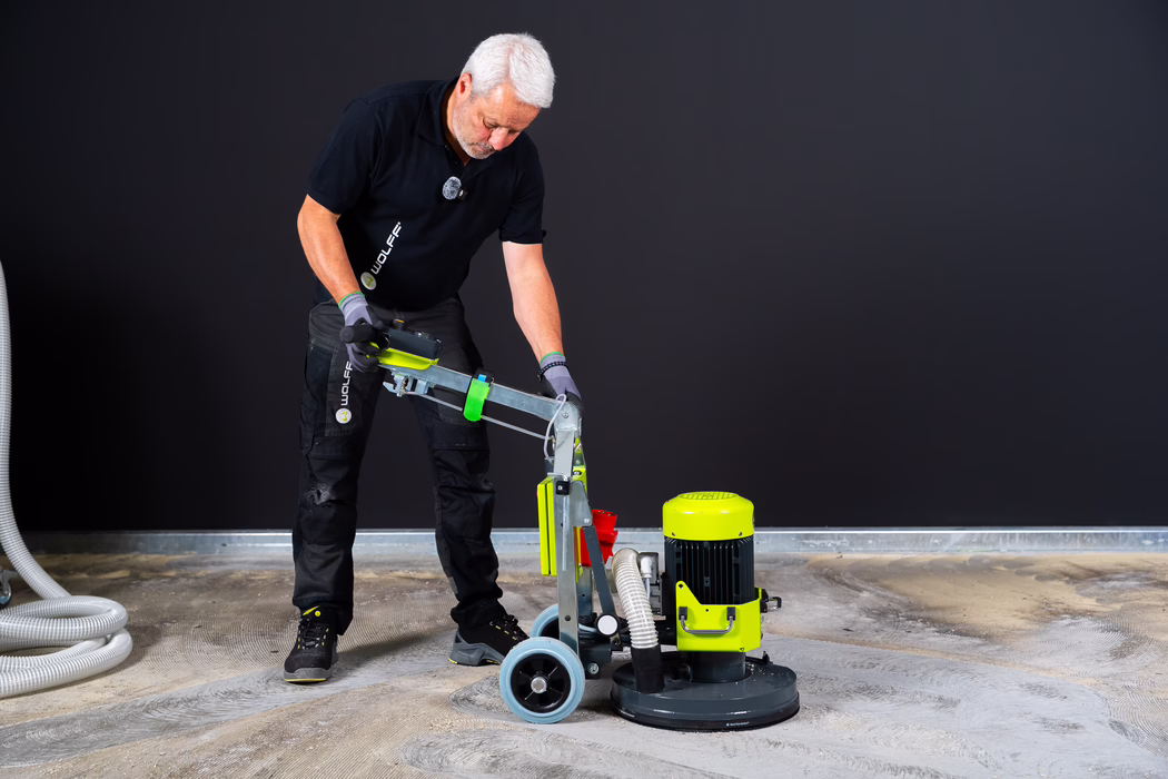 WOLFF_Application Image_NEO 330_183050_21_2026-08 A middle-aged man with short white hair wearing a black polo and work gloves operates a bright yellow-green floor sanding machine on a workshop floor. He leans forward to adjust the handle while guiding the machine; hoses and dust-extraction components are connected, and a dark wall forms the background.