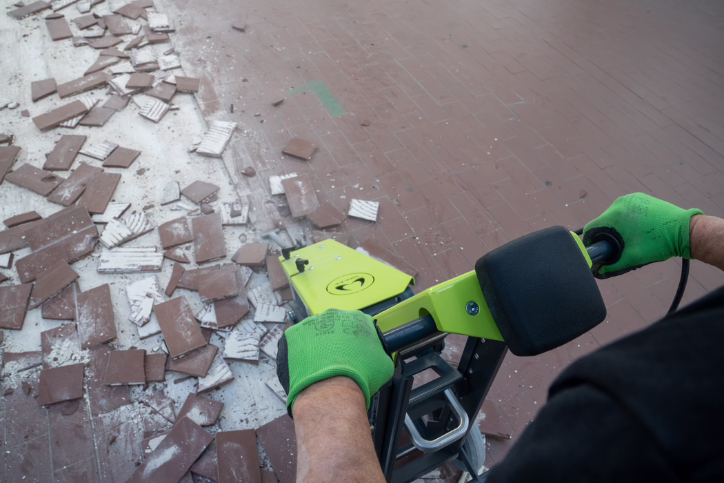 WOLFF_Application Images_RamHammer_Detail2_2022-11 Overhead view of a worker operating a bright green tile removal machine to scrape brown floor tiles; broken tile pieces and dust are scattered on the left side of the scene. The worker wears vivid green protective gloves and grips the machine handle, with a partially stripped tiled floor visible and the equipment seen in the lower right. Close-up perspective showing an active interior work task with debris and tools.