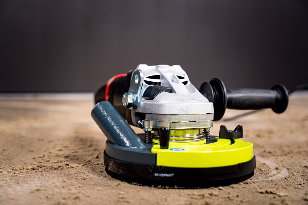 WOLFF_Application Images_BS 180_2024-01 (11) Close-up of a handheld angle grinder resting on a dusty concrete floor. The image shows the metallic motor housing with ventilation slots, a lime-green protective guard around the rotating head, a black auxiliary handle extending to the right, and a textured base covered in fine sand and dust. Shot with shallow depth of field, the background is blurred to emphasize the tool's metal details, bolts and signs of use.