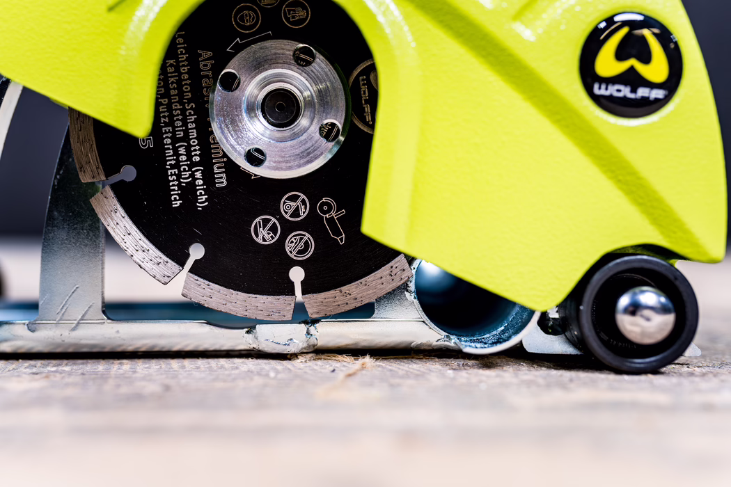 WOLFF_Application Image_18V EF135_Detail3_2022-11 Close-up of a circular diamond saw blade partially enclosed by a bright yellow protective guard bearing the 'Wolff' logo, resting on a rough surface. Visible are metal tooth segments, mounting holes in the blade, printed safety icons, and a small roller wheel at the base.