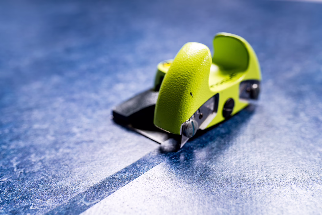 WOLFF_Application Image_Linocut_Detail4_2022-11 Close-up of a small bright green handheld paper cutter placed on a textured blue surface. The metal cutting blade and mechanism are visible in the foreground, while the plastic body shows a textured grip with a small black mark. Shallow depth of field blurs most of the background, emphasizing the cutter's shape, color and surface details.