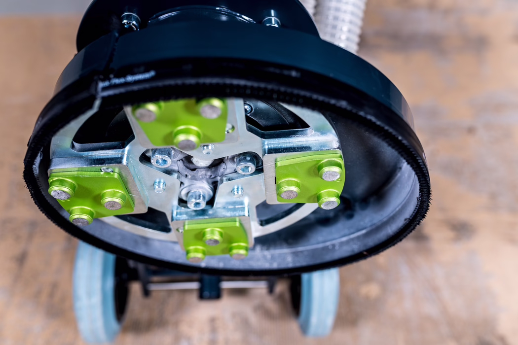 WOLFF_Application Image_Neo230_Detail12_2022-11 Close-up view of the underside of a floor grinding or polishing machine: circular metal base with four lime-green abrasive holders, visible bolts and central coupling, a black rubber skirt around the edge, and two small caster wheels with a wooden floor blurred in the background.