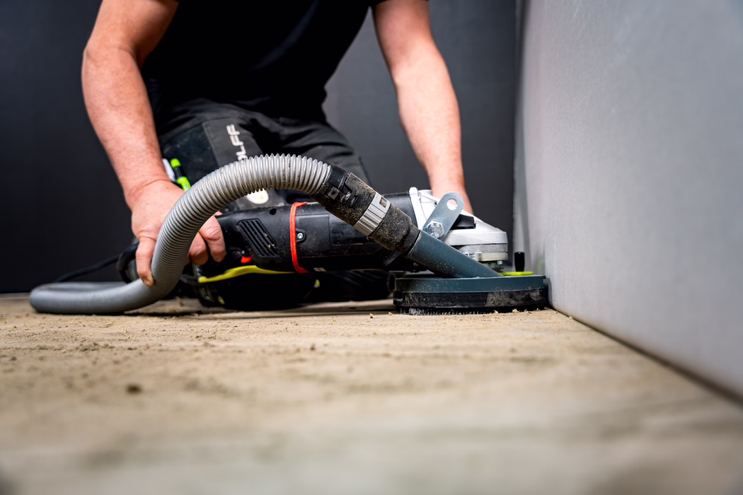 WOLFF_Application Images_BS 180_2024-01 (19) Close-up of a worker using a handheld electric floor sander along the base of an interior wall. The image shows forearms and hands gripping the sanding machine and the flexible dust-extraction hose attached to it. The sanding disc is pressed against a rough floor near the wall edge, producing fine dust. The worker wears dark work clothing and is crouched low to reach the floor; the background includes a dark wall and a smooth light-gray adjacent wall. The low-angle composition focuses on the tool and floor surface, with dust and textured floor in the foreground slightly out of focus.