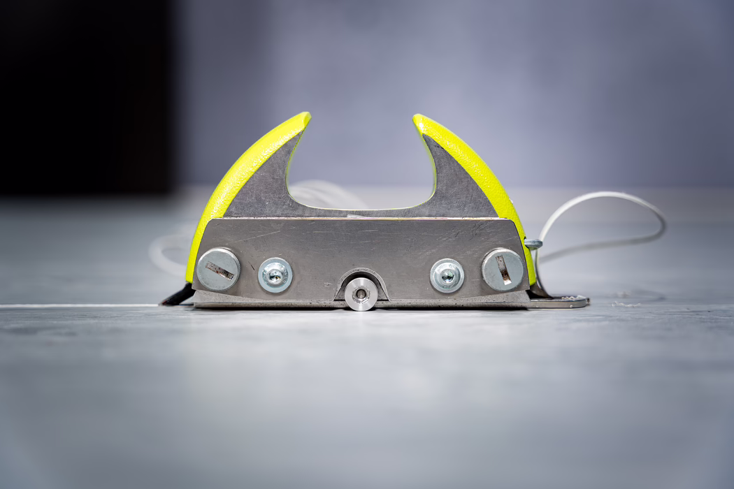 WOLFF_Application Image_Groover_2024-01 (6) Close-up low-angle photograph of a small metal floor-mounted guide or roller centered in the frame. Two curved, yellow-painted metal guards rise on either side of a flat brushed-steel base plate; the base shows several screws and a small central wheel or roller visible at the bottom center. A white cable or cord loops to the right and fades out of focus. The metal surfaces display fine scratches and signs of use, while the yellow paint has a slightly textured finish. The background is softly blurred cool gray with shallow depth of field, creating a minimalist industrial atmosphere. Diffuse, even lighting highlights the metallic textures and the bright yellow accents, emphasizing the object's functional, mechanical character.