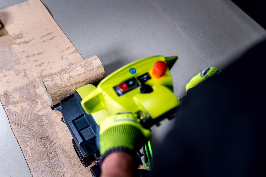 WOLFF_Application Image_Turbo_Detail2_2022-11 Close-up of a bright green floor sanding machine in use on a wooden surface: a person wearing a neon safety glove grips the handle while the rotating sanding drum smooths a strip of wood or old adhesive-covered flooring. Visible on the machine are control buttons including a red emergency stop, and a partially unrolled sanding belt; soft overhead light highlights dust and texture. The photo is framed from above, focusing on the tool and the worked area, with the operator only partly visible.