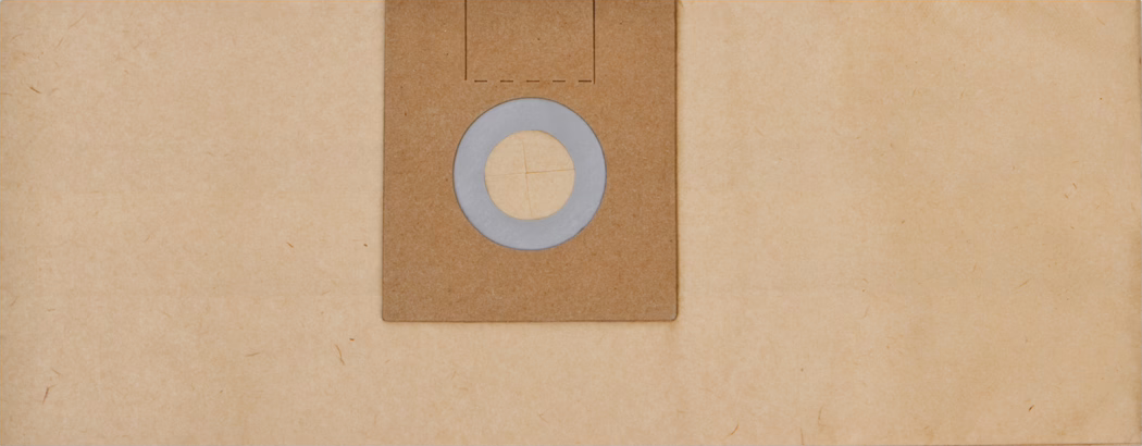 Close-up, centrally framed view of a square brown cardboard vacuum cleaner bag collar on a light beige textured paper background. The cardboard piece features a circular grey rubber or foam gasket surrounding a round opening with four inward-cut flaps. The surface shows subtle paper fibers, soft even lighting and muted warm tones, producing a minimal, tactile composition with slight edge wear and small creases.