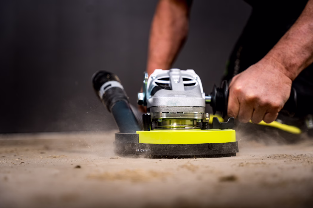 WOLFF_Application Images_BS 180_2024-01 (18) Close-up of a worker operating a handheld floor grinding machine on a concrete surface; the yellow abrasive head spins and raises dust while strong hands grip the tool's handles.