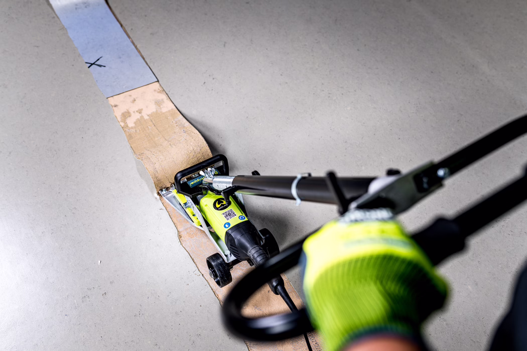 WOLFF_Application Image_Durostripper_Detail2_2022-11 Close-up view of a worker operating a small electric wheeled floor scraper over a strip of old adhesive on a grey concrete floor. A neon-yellow scraper tool is pushed along the adhesive; the gloved hand in a high-visibility green glove, the tool handle, power cord and small wheels are visible in the foreground. The scraped brown adhesive strip runs vertically through the frame and there are dust particles and scuff marks on the surrounding floor, suggesting a maintenance or renovation task removing floor covering residue.