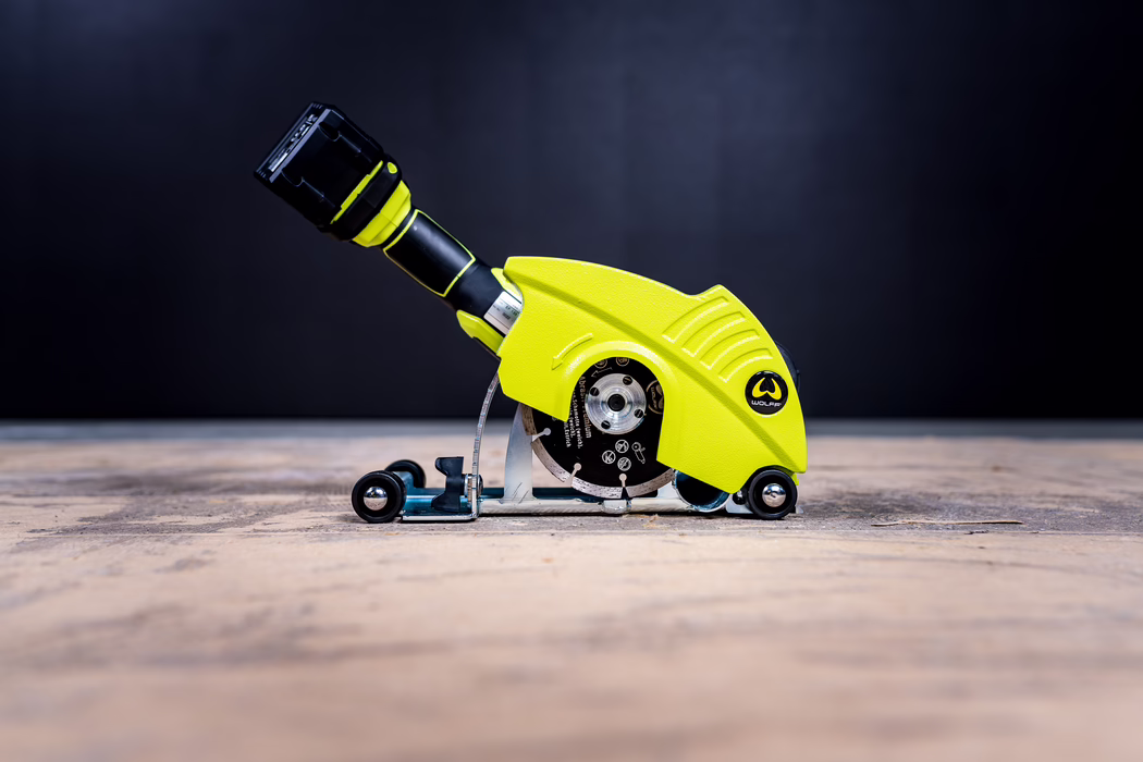 WOLFF_Application Image_18V EF135_Detail1_2022-11 Compact cordless mini circular saw with bright lime-yellow protective housing and a small diamond cutting disc, positioned on a wooden floor against a dark background; visible guide wheels and an ergonomic black handle with removable battery pack; designed for precise, portable cutting of tiles and flooring. Side view at low angle, studio-style lighting and high contrast.