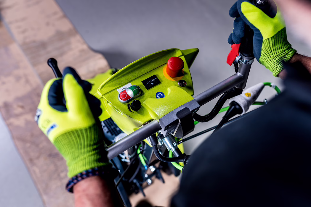WOLFF_Application Image_Biturbo_Detail4_2022-11 Close-up of a bright green machine control panel with two gloved hands on the handles. Visible are a red emergency stop button, a red-and-green start/stop switch, a small digital meter, cables and a metal handlebar. The operator is wearing high-visibility green work gloves and stands above a wooden floor or pallet. The image focuses on controls and safety features in an industrial or maintenance setting.