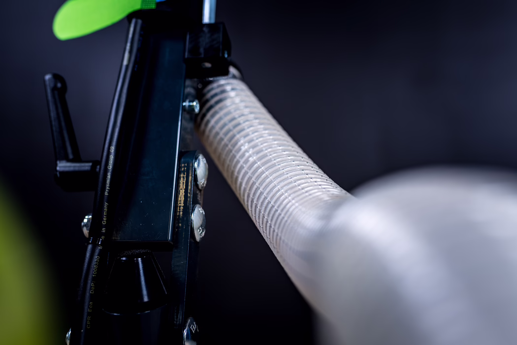 WOLFF_Application Image_Neo230_Detail2_2022-11 Close-up photograph of a mechanical component: a black metal bracket with visible bolts beside a textured white corrugated hose running diagonally into the foreground. A small green element appears at the top left. The background is dark and out of focus with cool, desaturated tones and shallow depth of field.