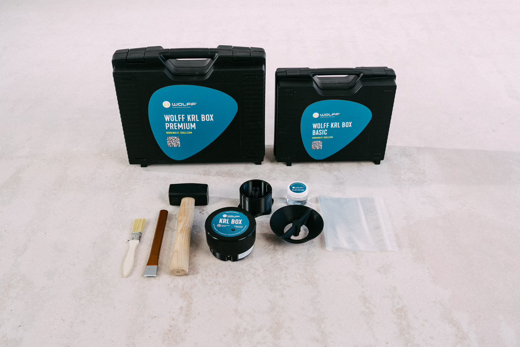WOLFF_Campaign Images_KRL Box_2025-08 (34) Two black plastic tool cases with teal-blue triangular labels reading 'WOLFF KRL BOX PREMIUM' and 'WOLFF KRL BOX BASIC' stand on a pale tiled floor. In front of them are arranged tools and accessories including a rubber mallet with a wooden handle, a small paint brush, a chisel, a round container labeled 'KRL BOX', a small jar, a plastic funnel, a black cylindrical part and a clear resealable plastic bag. The scene is well-lit with a neutral, minimal background and the items are organized in a neat row for presentation.