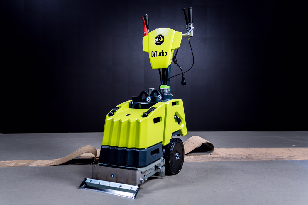 WOLFF_Application Image_Biturbo_Detail1_2022-11 A compact lime-green industrial floor machine labeled BiTurbo with black and metallic accents. The device has a tall handle with controls and an electrical cord, large rear wheels, and a metal front scraper or squeegee attachment. It stands on a concrete floor with a partially removed strip of flooring behind it; the dark studio-like background and directional lighting emphasize the textured plastic housing and visible mechanical details.