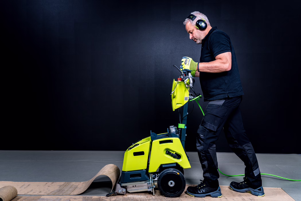 WOLFF_Application Image_Biturbo_Detail5_2022-11 A worker wearing ear protection and gloves operates a bright yellow-green floor stripping machine to remove an old carpet, guiding the equipment across an indoor concrete floor against a dark backdrop.