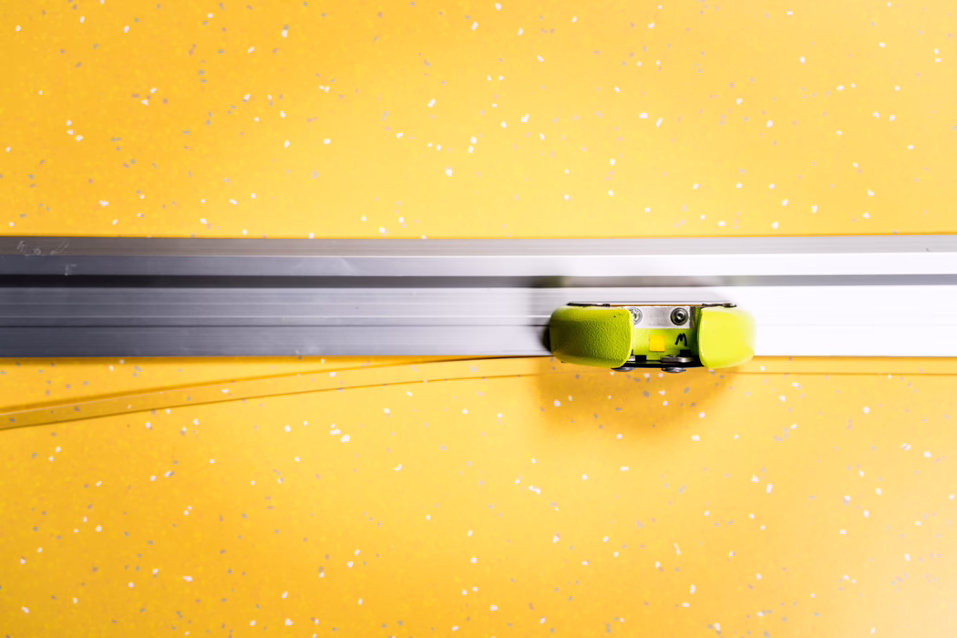 WOLFF_Application Image_Railcut_Detail3_2022-11 A close-up of a sliding door mechanism: a silver metal rail spans the frame with a small green roller carriage attached, set against a bright yellow speckled background.