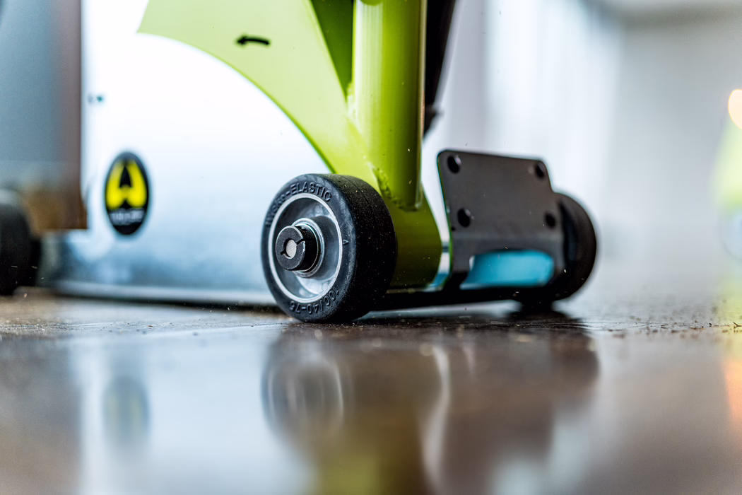 Low-angle close-up photograph of a small black rubber wheel mounted on a lime-green metal frame, resting on a polished wooden floor that reflects the wheel. The wheel features a metal hub with embossed markings; the green frame attaches to a gray metal base with a yellow-and-black warning sticker. The scene has a shallow depth of field, visible dust particles and scratches on the floor, and a softly blurred interior background.