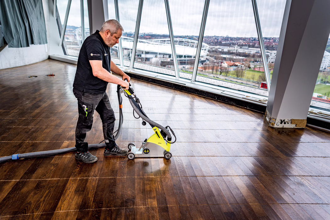 A worker is sanding a large dark wooden floor with a yellow floor sander inside a bright room with tall windows overlooking a stadium; he wears dark work clothes and safety shoes.