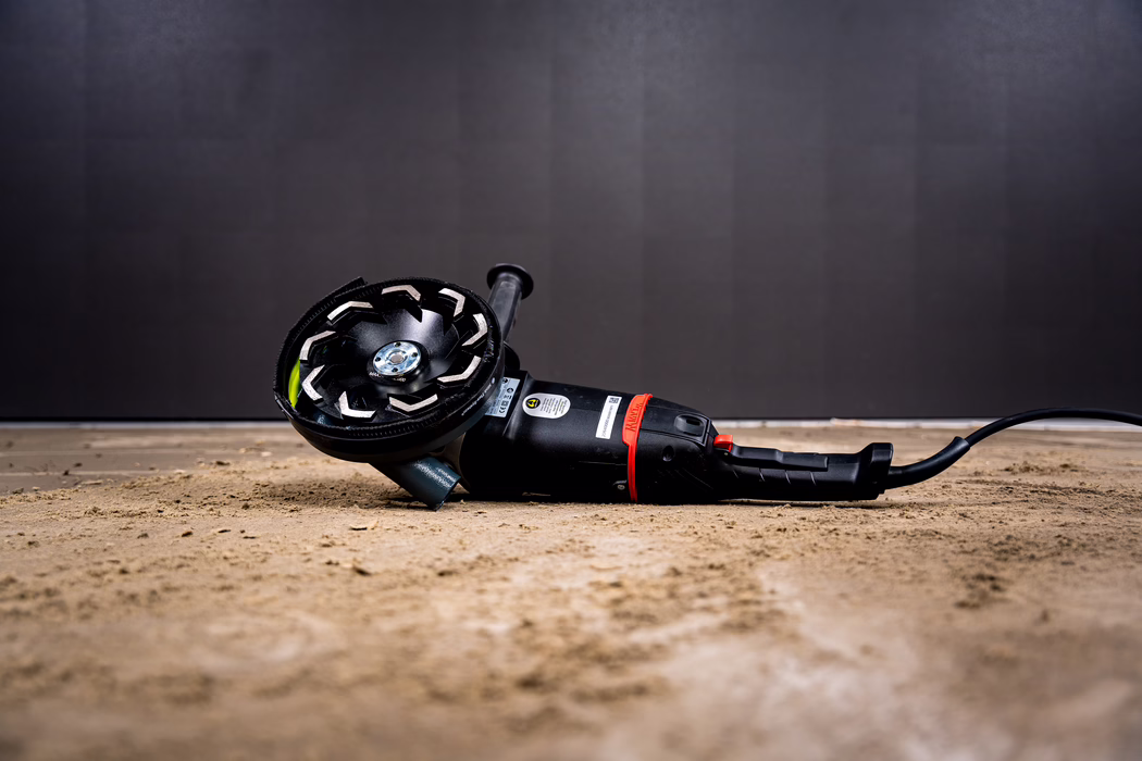 WOLFF_Application Images_BS 180_2024-01 (1) A close-up photo of a black electric angle grinder lying on a dusty concrete floor. The tool is positioned horizontally with the grinding disc facing the camera; the disc has segmented abrasive teeth and a metal center nut. The handle and power cord extend to the right; the body shows a red safety switch and a manufacturer's sticker. The background is a dark, out-of-focus wall, creating a low-key industrial atmosphere; fine dust and small debris are scattered on the floor around the tool.