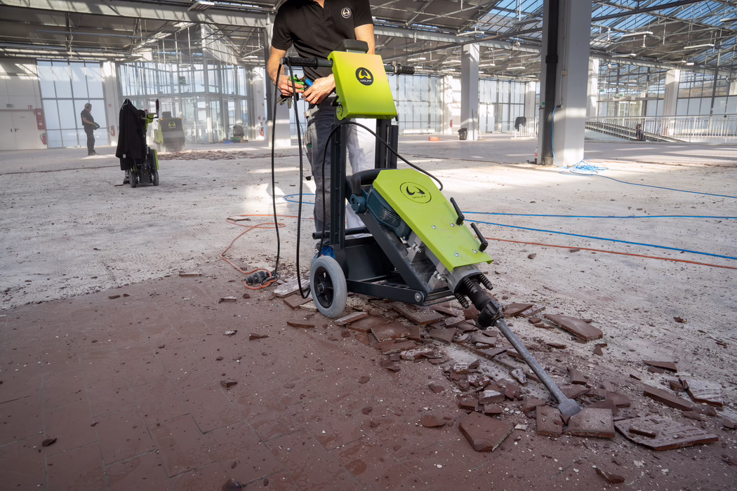 WOLFF_Application Images_RamHammer_Detail1_2022-11 A maintenance worker in a large, sunlit industrial hall operates a green floor-removal machine that breaks and lifts old red-brown tiles; shards and dust are scattered across the concrete floor. The worker wears a black polo and light work pants; only the torso and hands are visible as he holds the machine's controls. The compact wheeled machine has a bright lime-green casing with a logo and a long chisel-like attachment that penetrates the tile surface. In the background another worker with hearing protection stands near equipment; large windows and steel roof trusses let daylight stream into the high, open space. Coiled cables, power cords and construction debris are spread around, indicating an interior renovation or demolition of the floor.