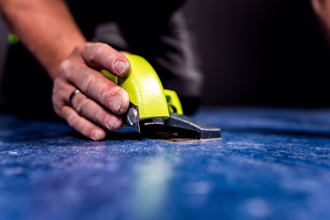 WOLFF_Application Image_Linocut_Detail1_2022-11 Close-up photograph with shallow depth of field showing a person's hand holding a bright lime-green handheld cutting tool with a small exposed blade. The hand, which has slightly worn fingernails and a simple ring on one finger, presses the cutter against a textured blue fabric or carpet laid on a flat surface. The metal blade and the textured painted surface of the tool are in sharp focus while the foreground and dark background are softly blurred, conveying concentrated manual work and precision cutting.
