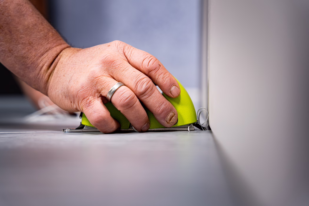 WOLFF_Application Image_Groover_2024-01 (10) Close-up of a person's hand holding a small bright green handheld scraper or sanding tool pressed against the base of a wall, shaving thin curled strips of material from a gray floor. The adult hand wears a silver wedding band on the ring finger and has short, slightly dirty nails. The tool has a lime-green plastic body and a metal base; the image is taken at a low angle with shallow depth of field, keeping the hand and tool in sharp focus while the background is softly blurred. Indoor, neutral lighting and muted tones. No faces or other identifying features are visible.