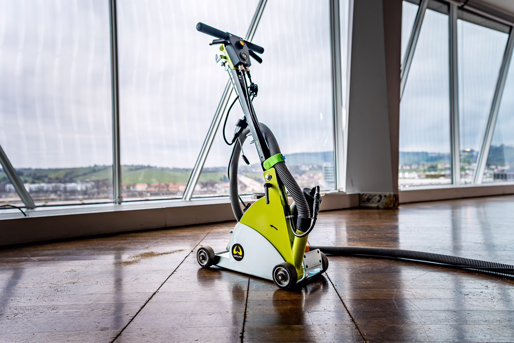 A lime-green floor sanding and polishing machine with hoses and small caster wheels stands on a glossy wooden floor in a bright room. The device features a curved metal body, a vertical handlebar with controls and a flexible vacuum hose coiled beside it. Large angled windows reveal a cloudy sky, a distant cityscape and rolling green hills beyond.