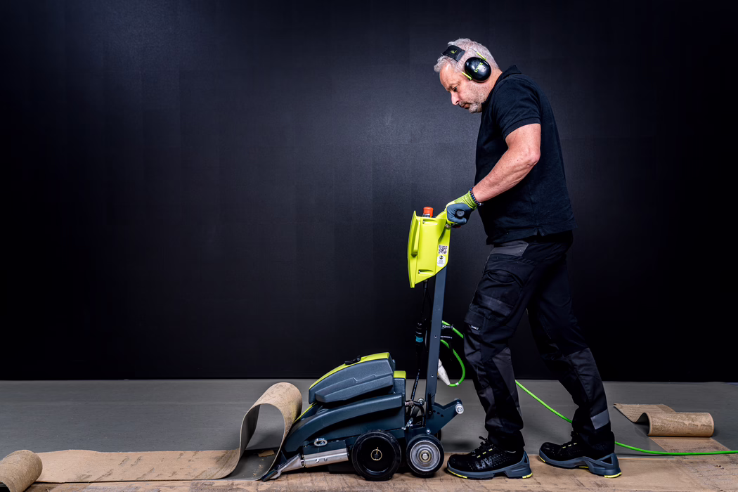 WOLFF_Application Image_Turbo_Detail1_2022-11 A professional tradesman operates a bright lime‑green floor stripping machine to remove old flooring. The man, wearing hearing protection earmuffs, work gloves, a black polo shirt, dark cargo trousers and safety shoes, leans forward to guide the machine along the floor. Several strips and rolls of the removed flooring lie rolled and partially peeled back around the machine; the device shows wheels, a handle with controls and a green power cable lying on the ground. The scene is lit with high‑contrast studio lighting against a plain dark backdrop, emphasizing the texture of the floor and the focused posture of the operator.