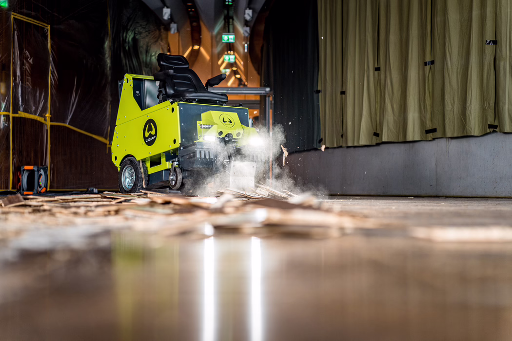 Low-angle, detailed view of a bright lime-green industrial floor-scraping machine working in a dim indoor corridor. The ride-on machine, branded with a circular 'WOLFF' logo on its side, has its front blade engaged and headlights on, kicking up a cloud of dust and wood splinters as it strips old wooden flooring. Debris and broken floorboards lie scattered across the polished surface in the foreground, with sparks of material and airborne particles frozen in midair by the camera. On the left side plastic sheeting and temporary scaffolding can be seen; on the right heavy beige curtains hang from the wall. Overhead exit signs and industrial lighting create a sense of depth down the corridor. The scene emphasizes motion, texture and contrast between the vivid machine and the muted, work-in-progress environment; no people are visible and the machine appears to be in active operation.