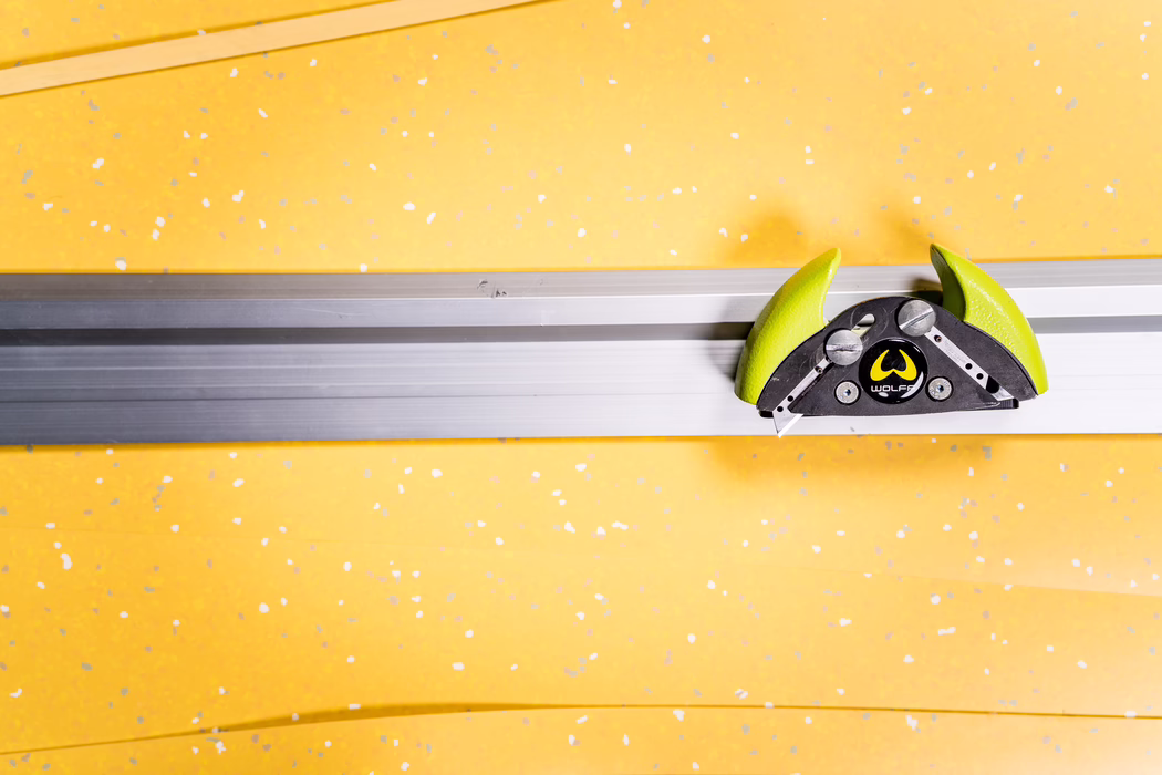 WOLFF_Application Image_Railcut_Detail2_2022-11 A close-up of a horizontal aluminum rail mounted against a bright yellow wall with white and gray speckles. On the right side a green and black Wolff-branded clamp or sliding tool is attached to the rail; its curved lime-green handles, black metal body, visible screws and a small retractable blade are clearly visible. The composition emphasizes strong horizontal lines, contrasting colors and a minimalist industrial aesthetic.