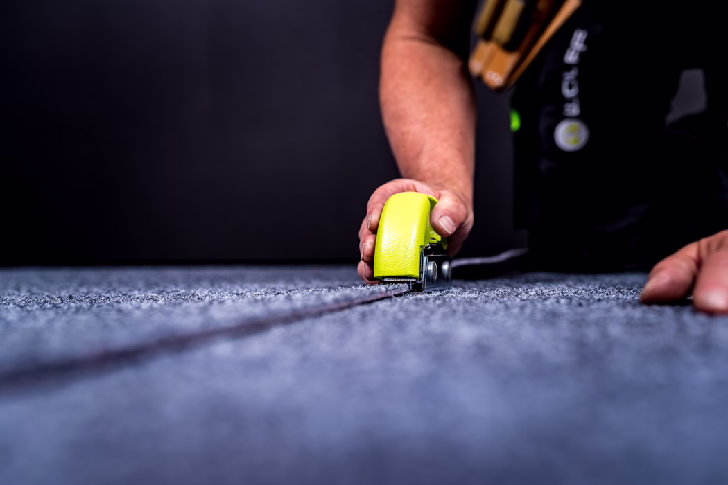 WOLFF_Application Image_GreenCut_Detail2_2022-11 Close-up of a person using a bright yellow seam roller to press and join a gray carpet seam. The foreground shows textured carpet fibers and a stitched seam line leading to the tool; the person's hand guides the roller while the other hand rests nearby. The background is dark and out of focus, emphasizing the tool and the working hands.