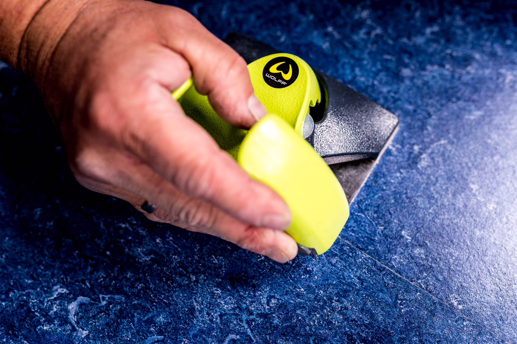 WOLFF_Application Image_Linocut_Detail3_2022-11 Close-up of a hand holding a bright yellow handheld tool with the 'Wolff' logo and a metal base, positioned on a dark blue textured stone or tile surface; focus on the fingers, the yellow plastic grip and the metal cutting or sanding edge.