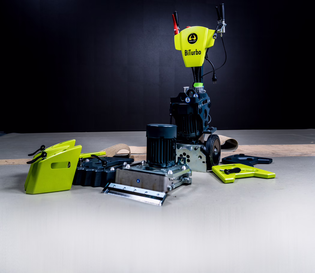 WOLFF_Application Image_Biturbo_Detail2_2022-11 Studio photograph of an industrial floor-preparation machine labeled 'BiTurbo' with a bright lime-green control housing and logo, two visible electric motor units, multiple detachable metal and plastic attachments, black power cables and wheels, placed on a concrete floor against a dark backdrop; the equipment appears arranged for presentation or inspection.