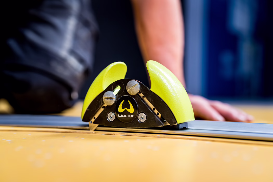 WOLFF_Application Image_Railcut_Detail5_2022-11 Close-up of a yellow-green WOLFF branded push block or clamp positioned on a metal track of a table saw. The textured plastic jaws, visible screws and the logo are in sharp focus, while the background with a person's arm and workshop surfaces is blurred; shallow depth of field and warm tones.