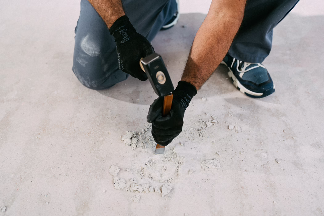 WOLFF_Campaign Images_KRL Box_2025-08 (53) Close-up view of a worker kneeling on a pale concrete floor while chiseling away old material. The image shows the worker's gloved hands gripping a chisel with a wooden handle and striking it with a small hammer to remove flakes of cement or plaster. The person's forearms are visible, wearing dark work gloves and gray work pants with dust marks; navy athletic shoes with white soles are partially visible. Fine dust and small rubble surround the chiseled area, and the soft even light emphasizes the textures of the concrete and tools. The shot conveys focused manual labor, renovation or repair work, and a careful, controlled technique.