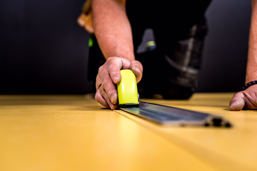 WOLFF_Application Image_Railcut_Detail1_2022-11 Close-up of a person's hand using a bright yellow seam roller to press a metal track onto a yellow floor panel; visible are the fingers, worn fingernails and parts of the forearms against a blurred dark background.