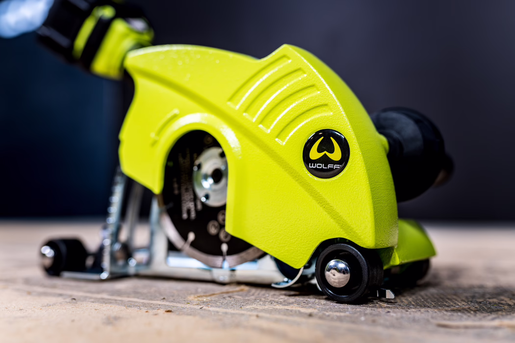 WOLFF_Application Image_18V EF135_Detail4_2022-11 Close-up of a compact handheld power saw with a bright lime-green plastic housing branded 'WOLFF'. The shot focuses on the textured guard and logo on the right side; a small circular metal blade is partially visible beneath the guard. Visible details include ventilation grooves in the housing, a metal baseplate, guide wheels and fasteners. The tool sits on a wooden surface with a shallow depth of field that softly blurs the dark background, while crisp lighting emphasizes the vivid color and surface texture.