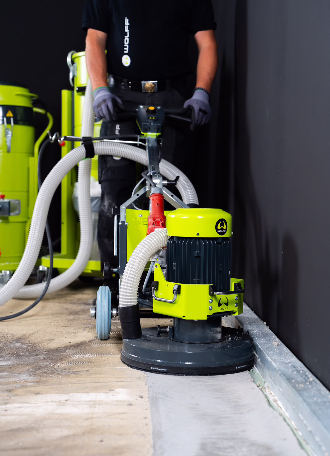WOLFF_Application Image_NEO 330_183050_19_2026-08 A worker operates a bright lime-green industrial floor grinding machine next to a dark wall. The close-up view shows the machine's rotating sanding head, large suction hoses connected to dust-extraction equipment, rubber wheels and a wheeled handle that the operator holds with gloved hands. The wooden floor shows sanding marks and an area of smoothed concrete where the grinder has removed the surface. The worker wears a black shirt and protective gloves; the face is not visible. The setting appears to be an indoor renovation or construction site.