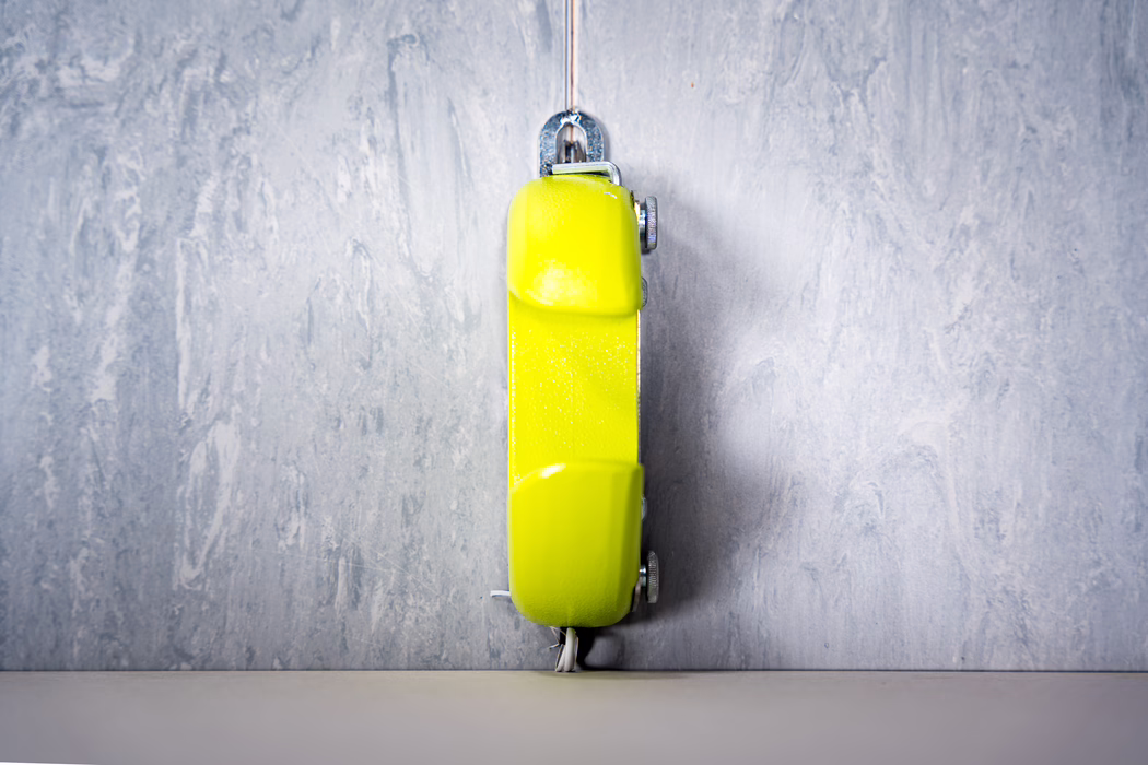 WOLFF_Application Image_Groover_2024-01 (11) Close-up of a bright yellow retractable tape measure hanging vertically against a grey textured concrete wall, shown from the side with the metal belt clip and winding knobs visible; the tape measure casts a soft shadow and a short length of white measuring tape peeks out at the bottom.