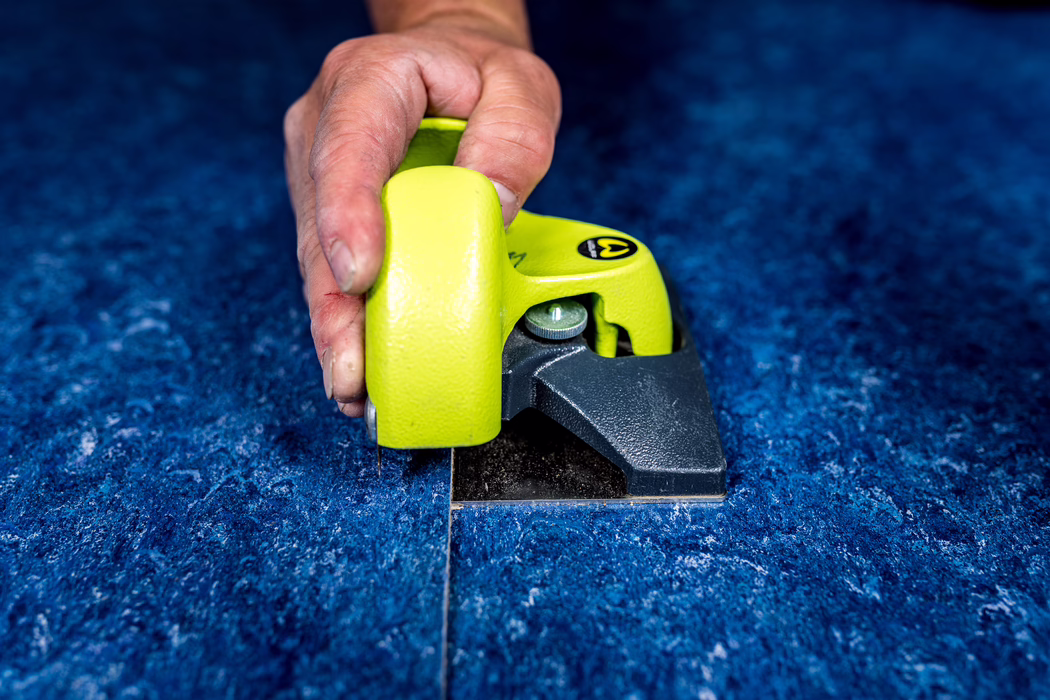 WOLFF_Application Image_Linocut_Detail2_2022-11 Close-up of a hand guiding a bright lime-green carpet trimming tool along the seam of a textured blue carpet; the tool's blade cuts a straight line while the person holds it firmly.