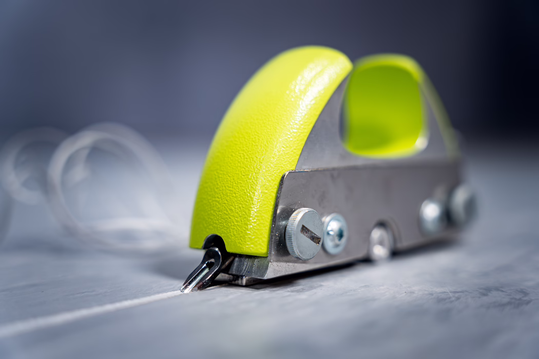 WOLFF_Application Image_Groover_2024-01 (7) Close-up of a small handheld metal tool with a curved, bright lime-green painted handle and visible screws. The metal tip or blade at the front is pressed against a light gray wooden surface, leaving a fine scored line. Soft studio lighting highlights the textured paint and metallic reflections while the background fades into a smooth blue-gray bokeh. The composition emphasizes texture, the color contrast between neon green and cool grays, and the precise mechanical details of the tool.
