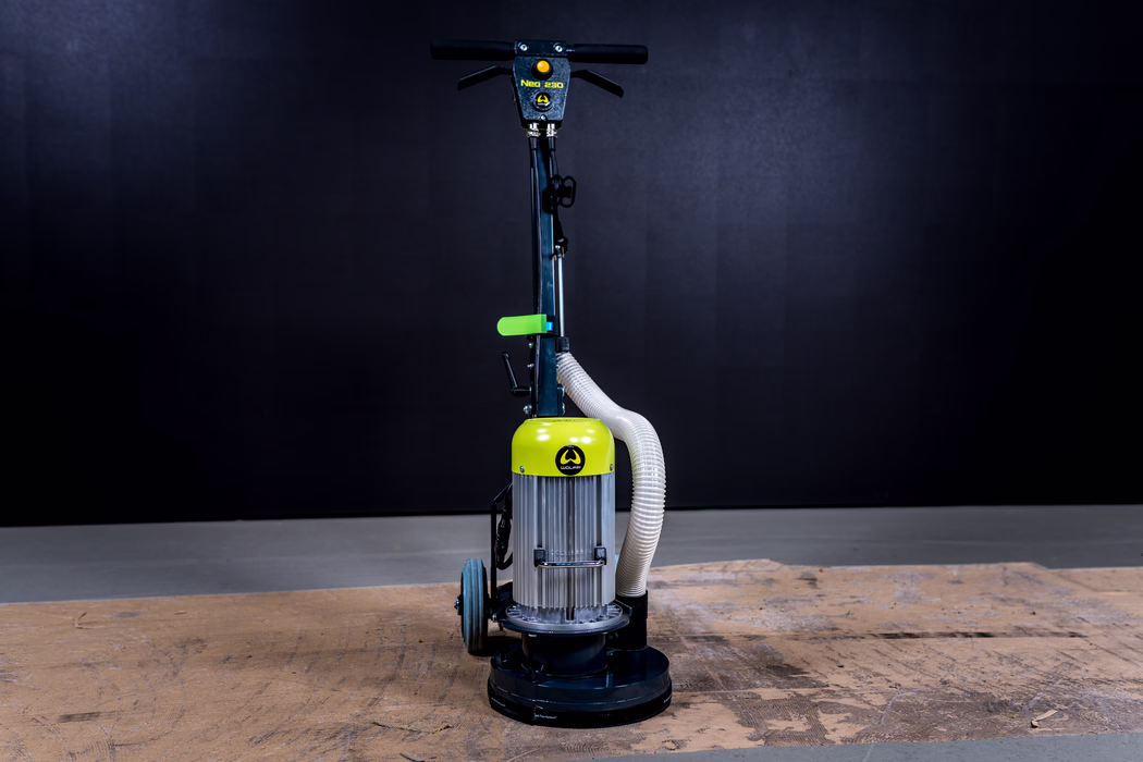 WOLFF_Application Image_Neo230_Main_2022-11 A single-disc floor sander/polisher photographed upright in a studio with a dark black background and worn wooden boards in the foreground. The machine has a cylindrical motor housing with a bright yellow-green top, vertical handlebar with a horizontal grip, two small transport wheels at the rear and a flexible white dust hose curving from the motor down to the sanding head. The round sanding base rests on the wood surface, and studio lighting highlights the machine's metallic textures, hose and controls while the background remains evenly dark.