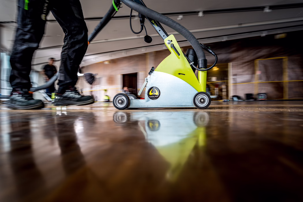 Low-angle close-up of a floor sanding machine in motion on a polished wooden floor inside an interior undergoing renovation. A lime-green and silver sanding unit with a visible 'WOLFF' logo and an attached flexible hose rolls on small wheels while a worker's legs in black work trousers and safety boots push it; only the legs and feet are visible. The glossy floor reflects the machine and lights, and the blurred background shows taped-off walls, equipment and other people, conveying an active construction or restoration scene.